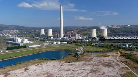 Steaming-thermal-power-plant-behind-the-lake-and-old-excavation-machine,-the-village-and-wind-turbines-in-the-background,-bright-and-sunny-afternoon