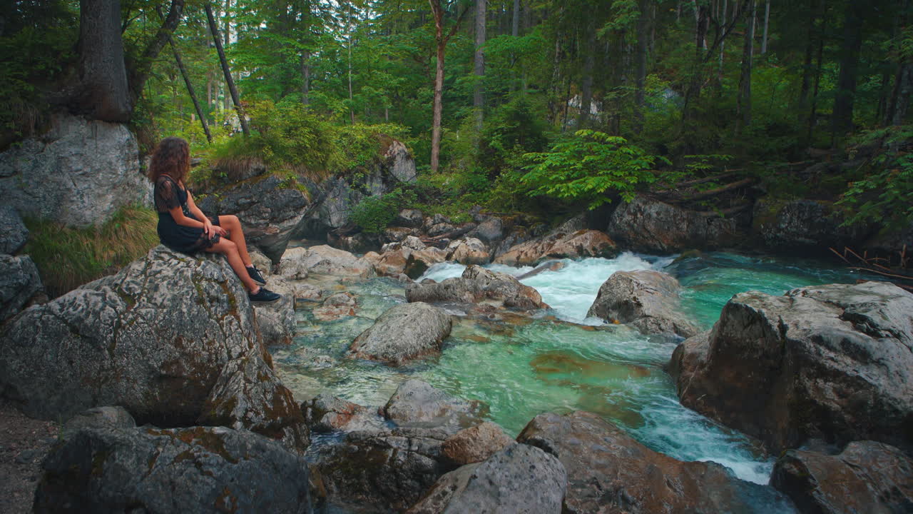 4K UHD Cinemagraph - seamless video loop of a young brunette woman sitting at a river at the scenic mountain lake Hintersee in Bavaria, Germany