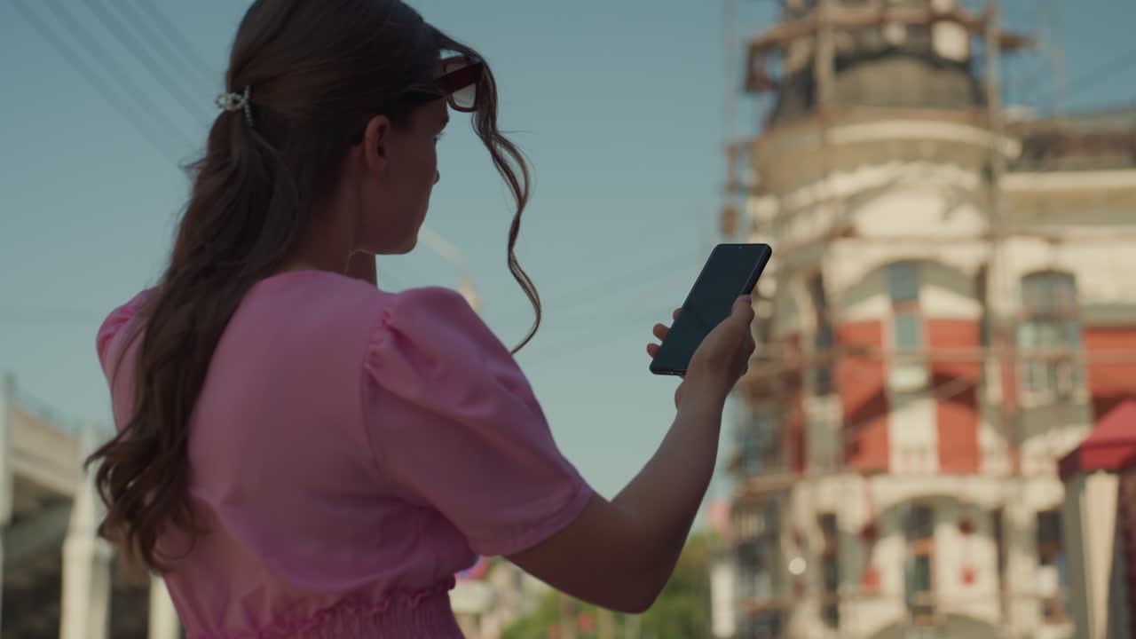 white woman pink dress holds phone while walking down sunny avenue, tapping screen to check messages, confident blogger stride in heels, candid street footage, warm light and lively downtown