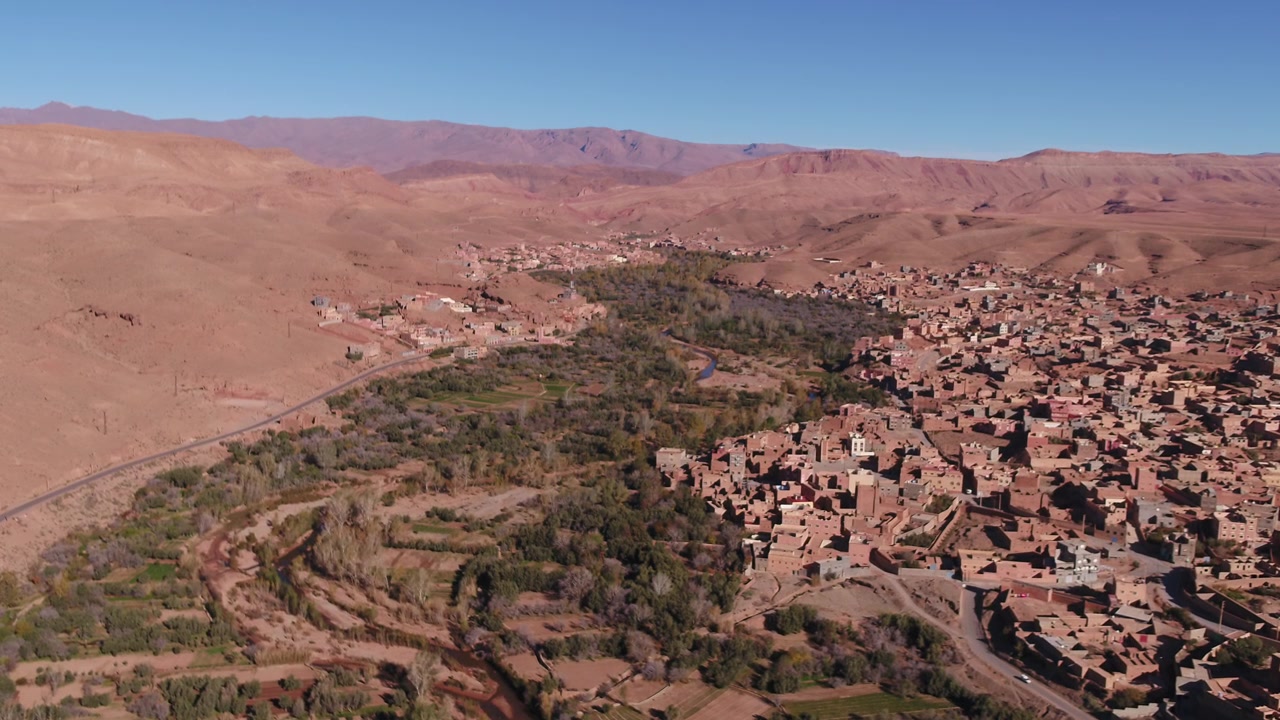 Veduta aerea obliqua ad alta quota da drone focalizzata sui densi edifici tradizionali in terra cruda e sulla disposizione urbana della città vecchia di Tinghir lungo il bordo della rigogliosa oasi in Marocco