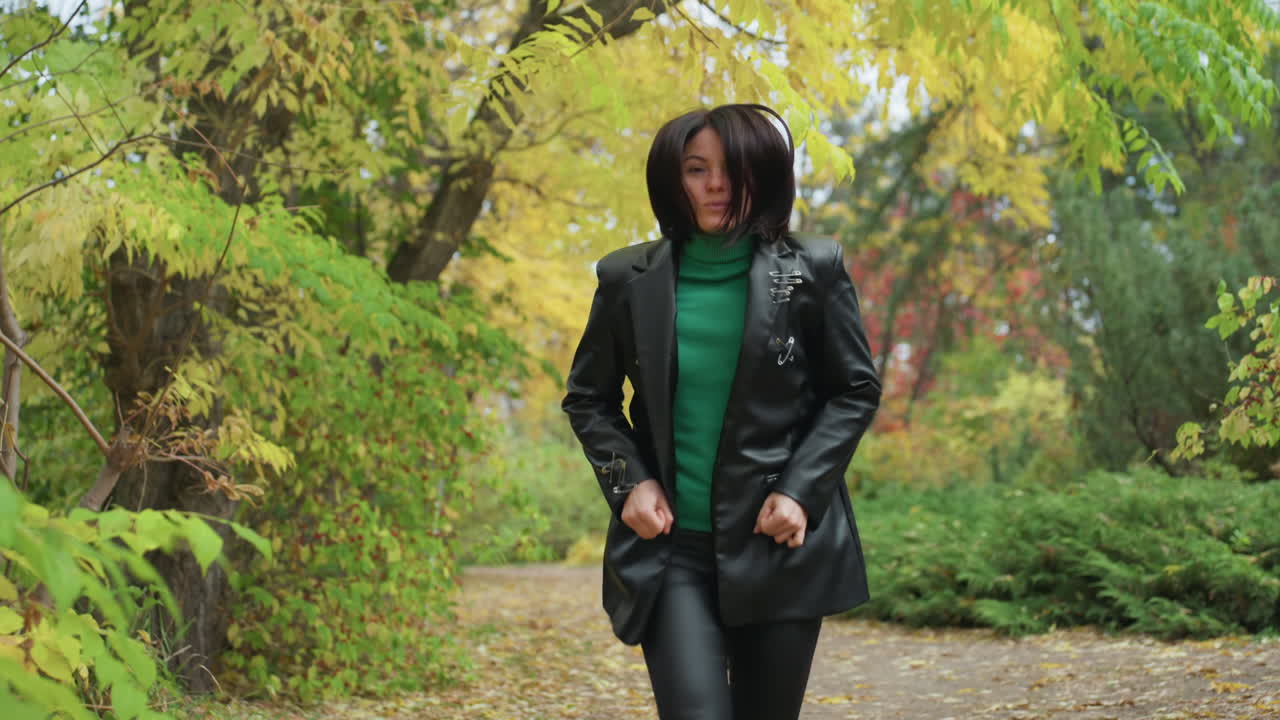 Donna di colore che cammina in una foresta autunnale con giacca di pelle, maglione verde e stivali, foglie che scricchiolano, passo sicuro, stile da musicista rock, espressione contemplativa, luce naturale, fogliame dorato, sentiero, primo piano e grandangolo