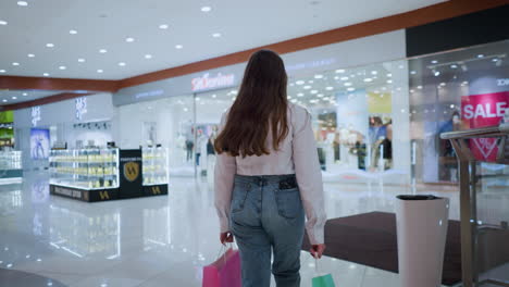 vista trasera de una mujer elegante con cabello largo caminando con confianza a través de un centro comercial moderno, ella lleva bolsas de compras en ambas manos mientras explora el ambiente comercial brillante y ocupado