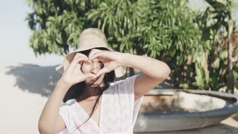 Portrait-of-happy-hispanic-woman-in-sunhat-making-heart-shape-with-hands-on-sunny-beach,-slow-motion