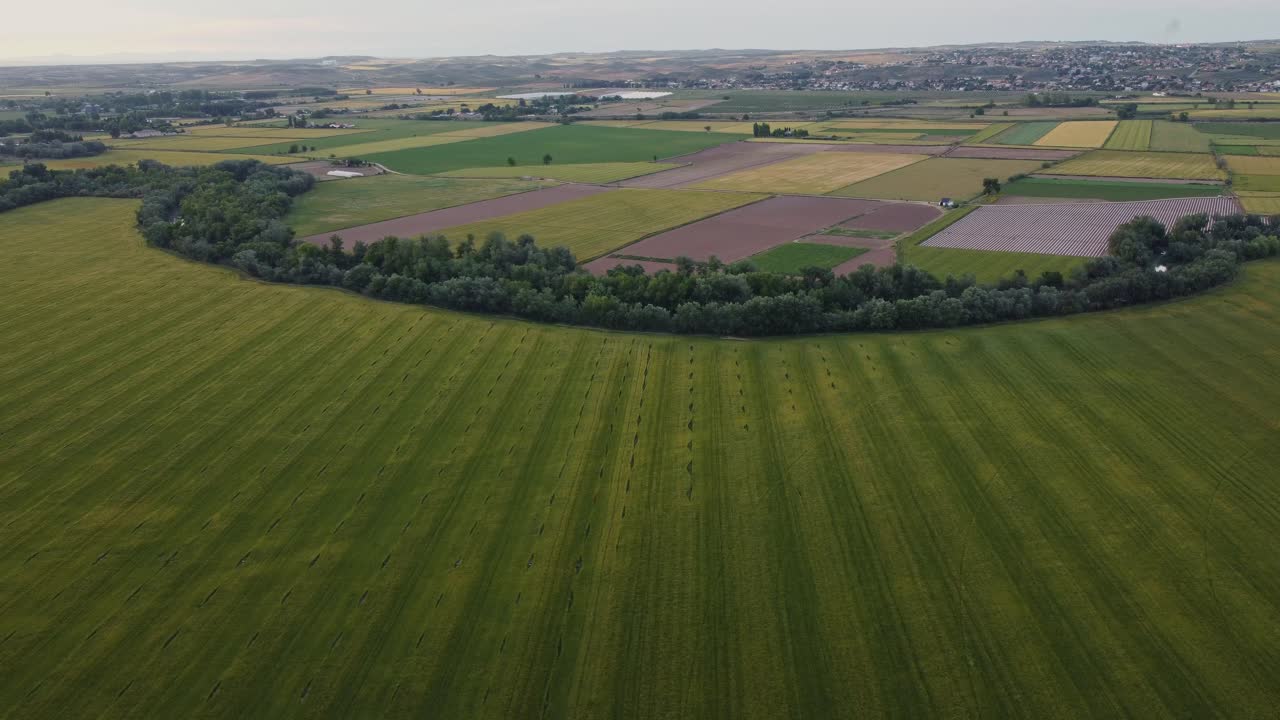 Drone shot flying over lush green fields and colorful farmlands near Aranjuez, Spain. Agricultural landscape with distant town and cloudy sky.
