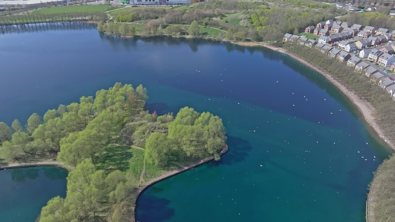 Drone footage over a Doncaster Lakeside in a forest next to a residential area. The lake features a small island. Located in Doncaster