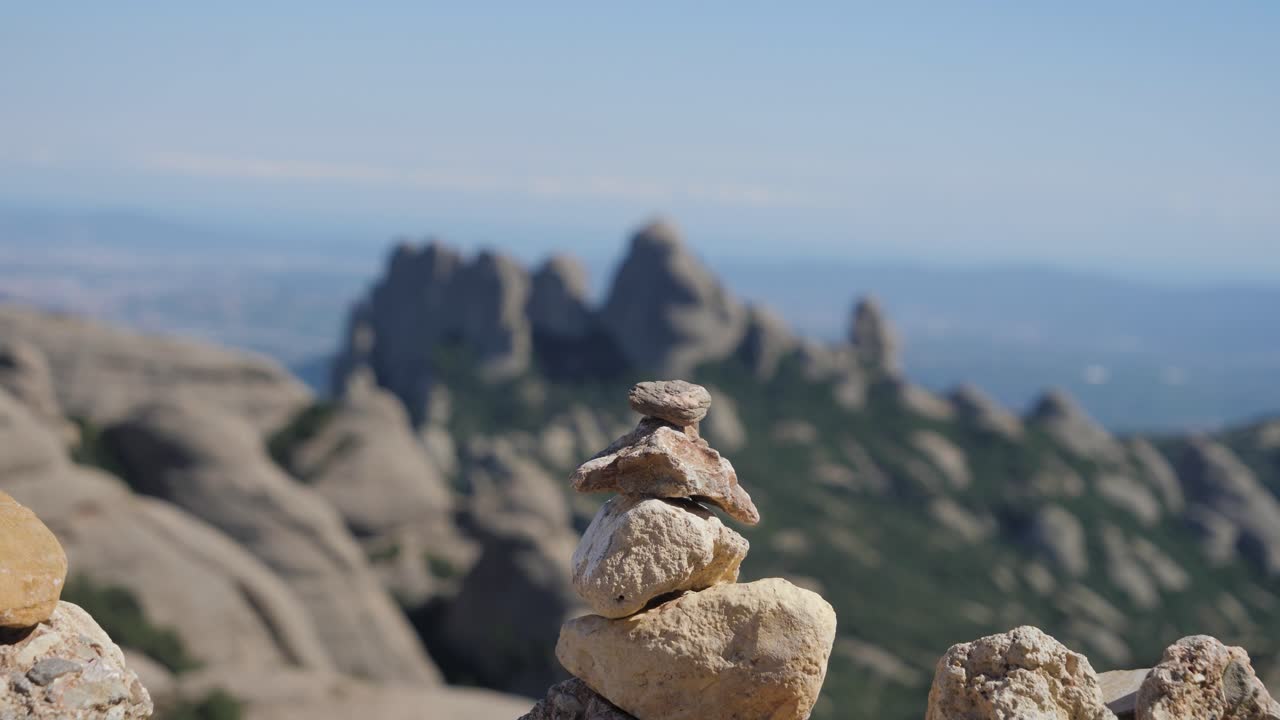 Et nærbillede af en hånd, der placerer en sten oven på en stabel sten ved et udsigtspunkt i bjergene i Montserrat, Spanien, med de takkede konglomerat-tinder synlige i den bløde baggrundsfokus.