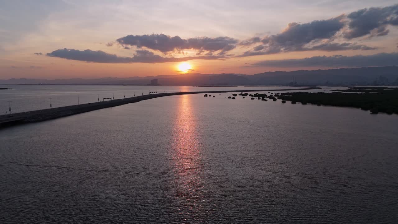 Cinematic drone view of the Davao Coastal Road causeway during a dramatic golden sunset. A modern highway curving along the shoreline with a bright sun reflection on the calm ocean water