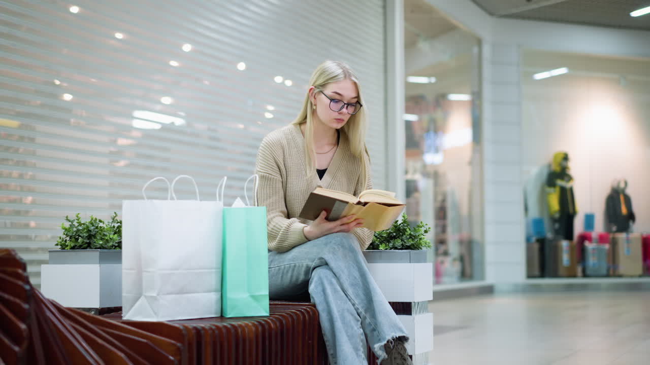 En rolig ung kvinde sidder i et lyst indkøbscenter med krydsede ben og læser en bog, mens indkøbsposer står tæt ved. En butik i indkøbscentret er synlig i det fjerne, hvilket skaber en afslappet og fredelig stemning.