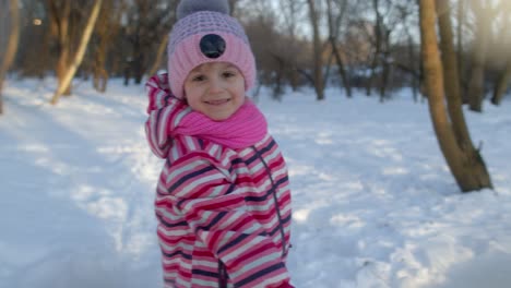 niña alegre lanzando bolas de nieve a la cámara, bromeando, sonriendo en el parque de invierno