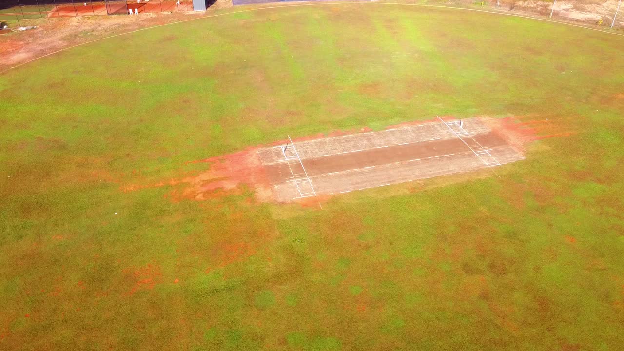 Ripresa aerea con drone dall'alto di un campo da cricket rurale. Il filmato mostra il wicket in terra battuta marrone, il campo esterno verde con chiazze di terra rossa e un semplice padiglione per gli spettatori.