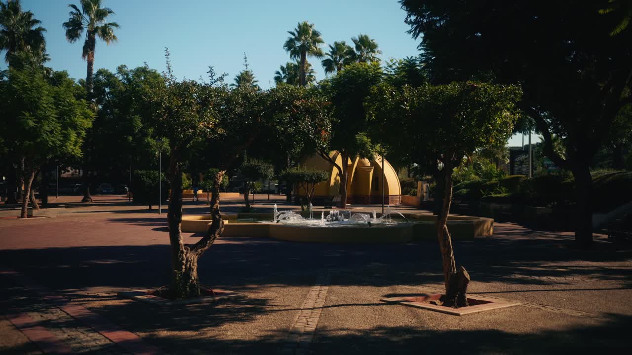 Piazza aperta con una fontana circolare e alberi sotto il sole splendente.