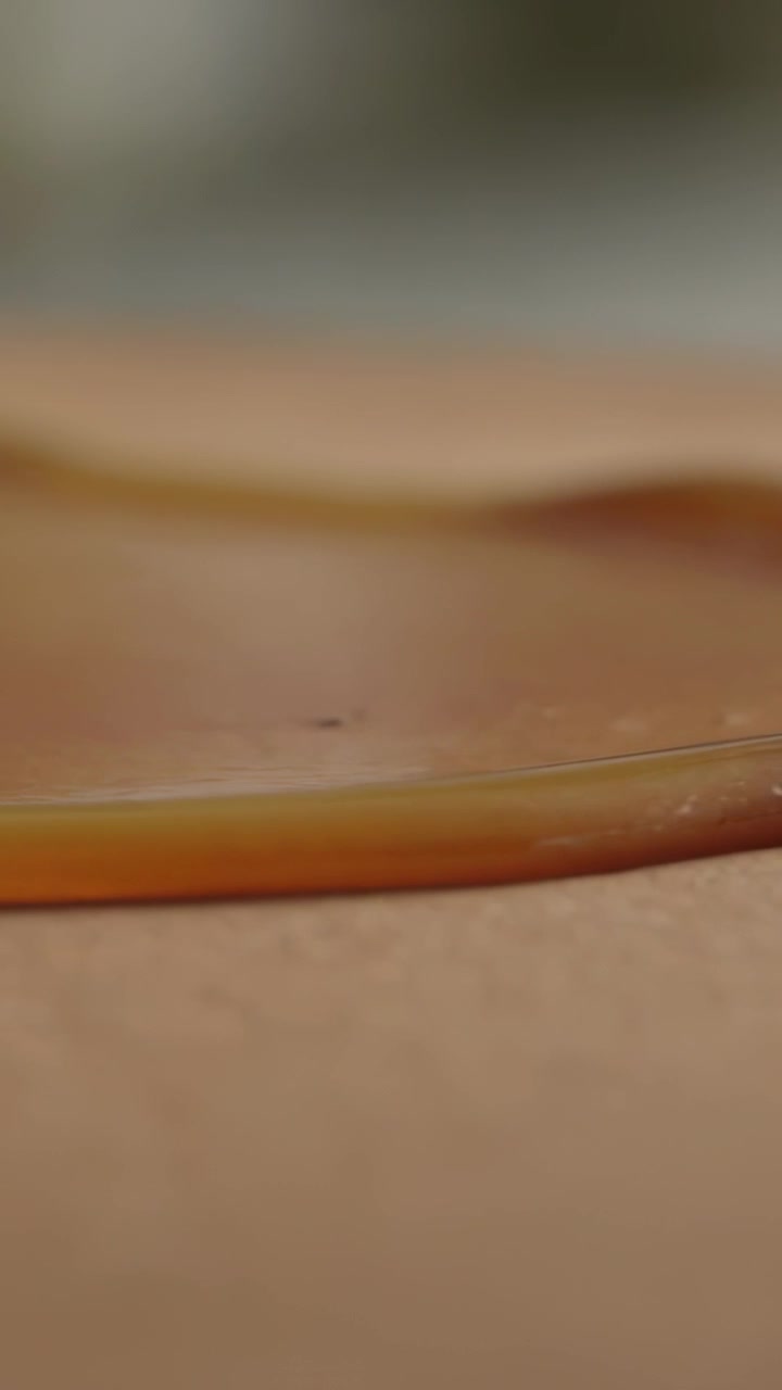 Video verticale: Pozza di liquido ambrato sottile che si assesta, levigando il bordo su un tavolo marrone chiaro, con particelle in movimento.