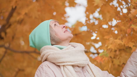 woman wearing mint beanie and warm scarf softly touches golden autumn leaf while gazing upward with peaceful expression, surrounded by vibrant orange foliage