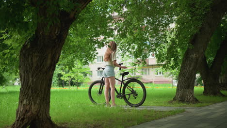 white lady approaches black bicycle parked on paved path, placing one glove on bike seat while wearing other, background features children playing in distance, tall trees, and residential buildings