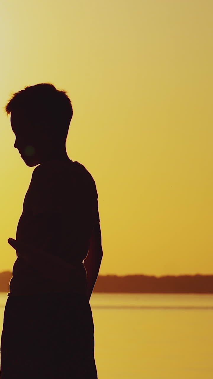 little kid holds origami of paper airplane in his hand and plays at sunset on the background of lake. A boy passionates about playing with his paper toy. Vertical video