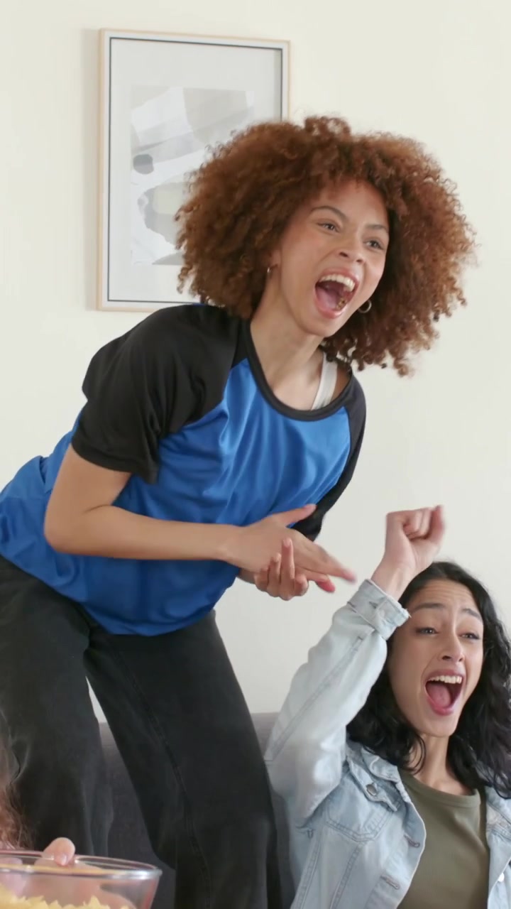 Vertical video: Cheering curly woman in blue top on sofa, clapping at offscreen play, chips bowl