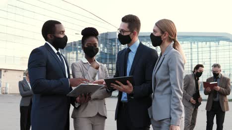 multiethnic group of business people in facial mask talking, using tablet and reading documents in the street