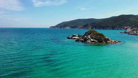 Small-rocky-island-surrounded-by-crystal-clear-blue-water-with-mountain-and-forest-in-the-background-shot-with-a-drone