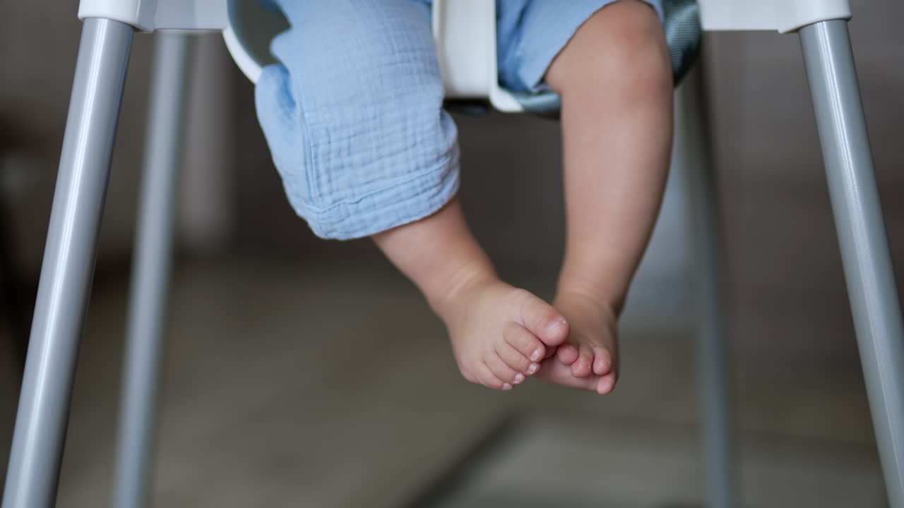 Lovely little baby feet hanging from a chair. Beautiful kid sitting at feeding table moves his tiny feet and toes. Close up.