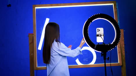 creative woman filming art. a young woman creates art while filming with a smartphone in a brightly lit blue studio space