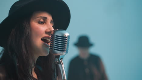 close-up view of a passionate singer in a black hat and leather jacket singing into a vintage microphone against a blue background. another person is blurred in the background