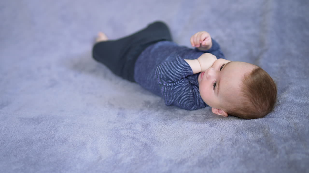 Calm little infant boy lying peacefully on his back. Beautiful child chewing his fingers and looking sideways. Blurred grey background.