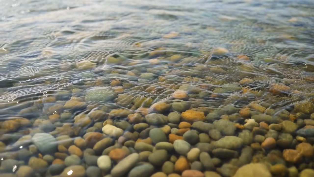 Primo piano di una spiaggia di ciottoli con acqua limpida, lievi increspature creano motivi scintillanti su pietre lisce e arrotondate illuminate dal sole