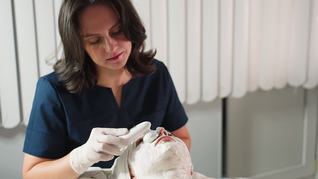 Beauty expert focused uses electric facial brush vibrating in circular motions to smooth thick cream mask on individual under bright spa light for deep skin rejuvenation and relaxation