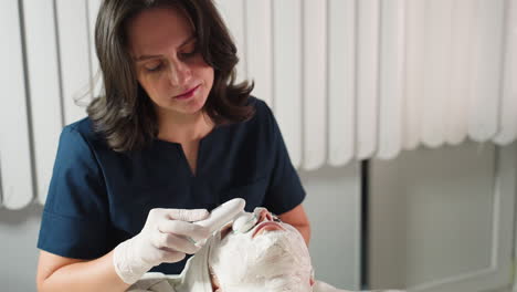 beauty expert focused uses electric facial brush vibrating in circular motions to smooth thick cream mask on individual under bright spa light for deep skin rejuvenation and relaxation