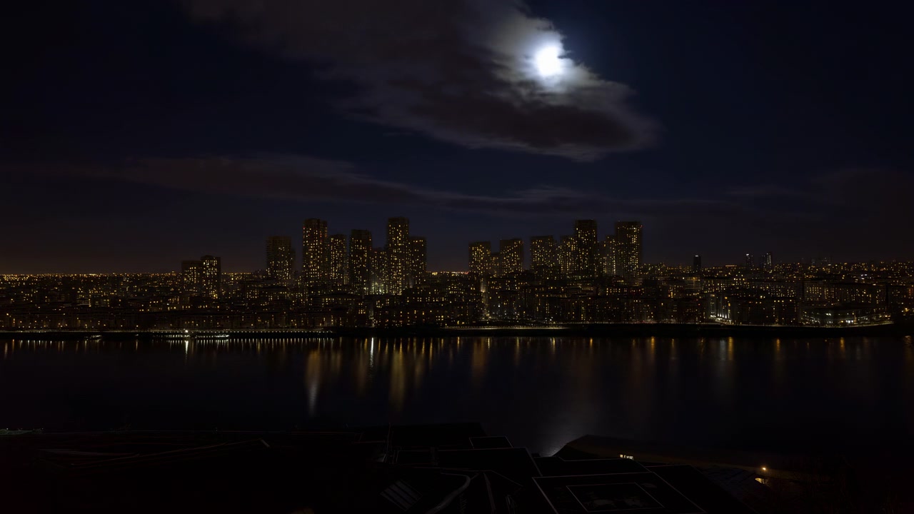 La vista notturna panoramica mostra densi grattacieli che brillano oltre l'acqua ferma, mentre le nuvole si spostano attorno alla luna luminosa sopra la testa e le strisce di luce della città scintillano in riflessi allungati
