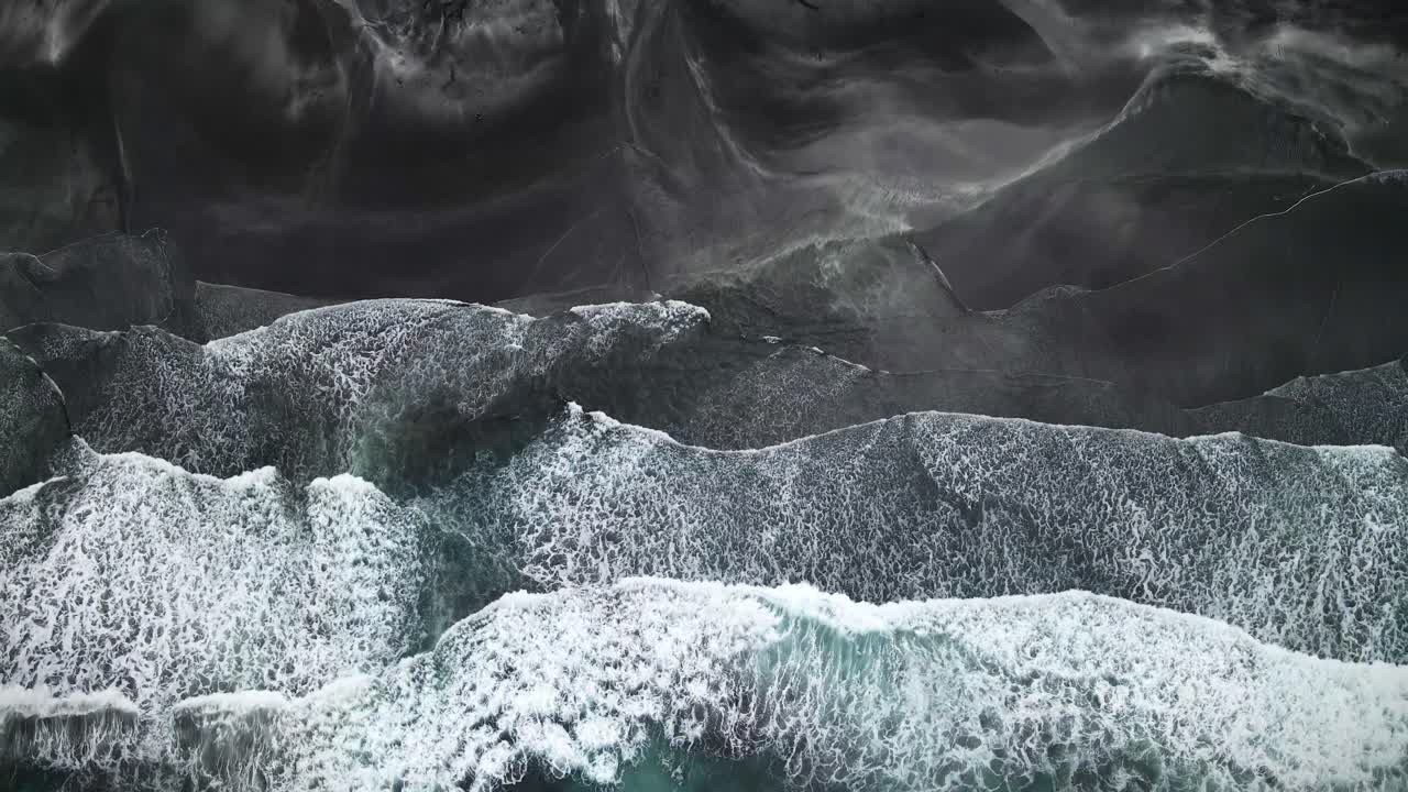 Aerial view of dramatic waves crashing against the dark rugged coastline of Tjørnuvík, Faroe Islands, highlighting striking contrasts between white surf and black terrain with vivid natural textures