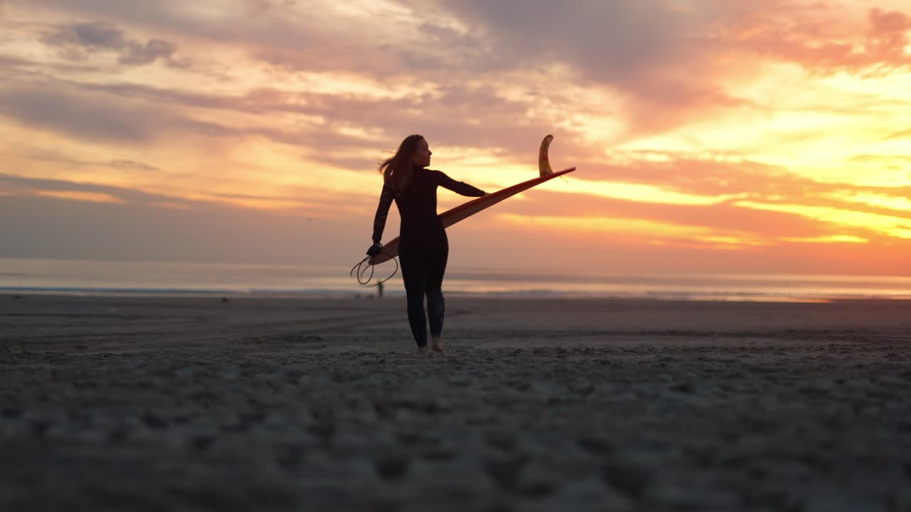 una fotografía amplia de una mujer surfista caminando por la playa con una tabla de surf al atardecer en costa da caparica, portugal
