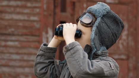 little red haired boy in hat and glasses looking in binoculars and dreaming to be an aviator