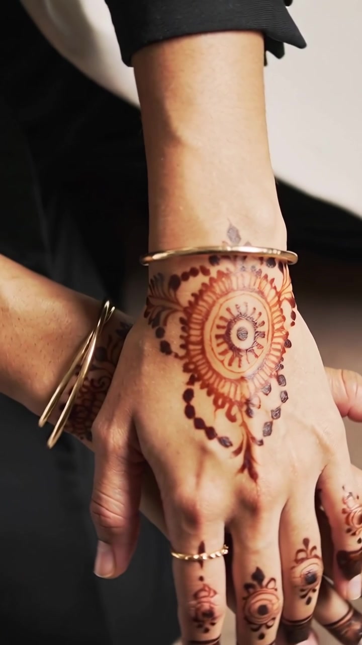 Video verticale: Zoom su mani con maniche scure che ruotano il polso, mostrando un mandala all'henné e braccialetti sul grembo.