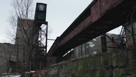 old, dilapidated industrial building with a rusted bridge connecting two sections, surrounded by overgrown vegetation under a cloudy sky