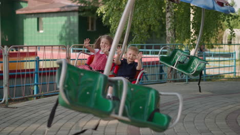 Happy-little-children-wave-hands-riding-carousel-in-park