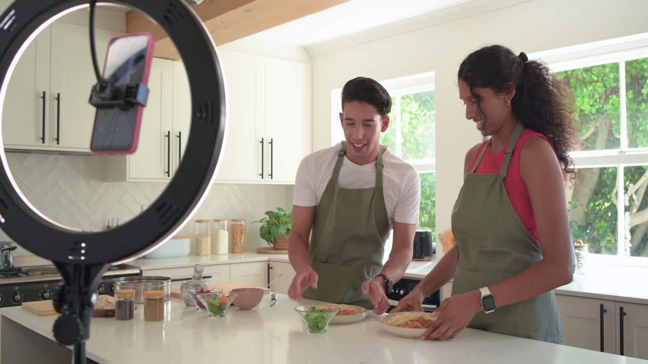 Coapresentadores afro-americanos com aventais verdes filmando com um celular com ring light, apresentando pratos em casa.