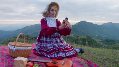 modelo rubio femenino en ropa tradicional anticuada sosteniendo un teléfono inteligente moderno tomando una selfie en la naturaleza mientras almuerza solo