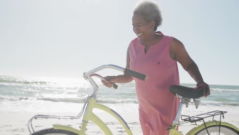 Happy-senior-african-american-woman-walking-with-bike-at-beach,-in-slow-motion