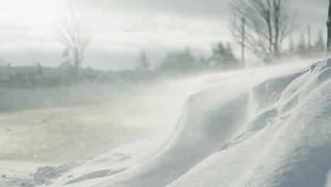 Wind-blowing-snow-across-a-road-side-snow-drift-in-slow-motion-on-a-country-road-in-bright-sunshine,-ending-in-a-whiteout