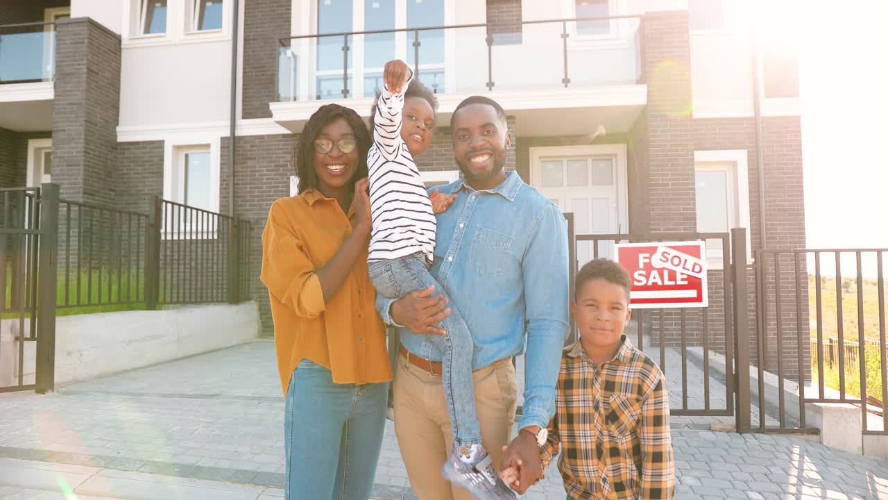 Portrait of happy African American family with small children standing at new house at suburb and showing key to camera