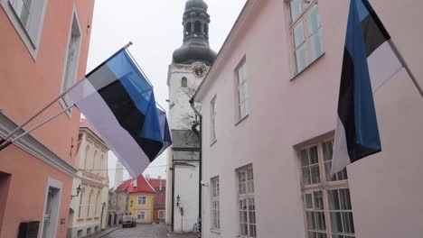 estonian-flags-hanging-on-pastel-building-with-church-in-the-background-with-grey-sky-in-Tallinn,-still-shot