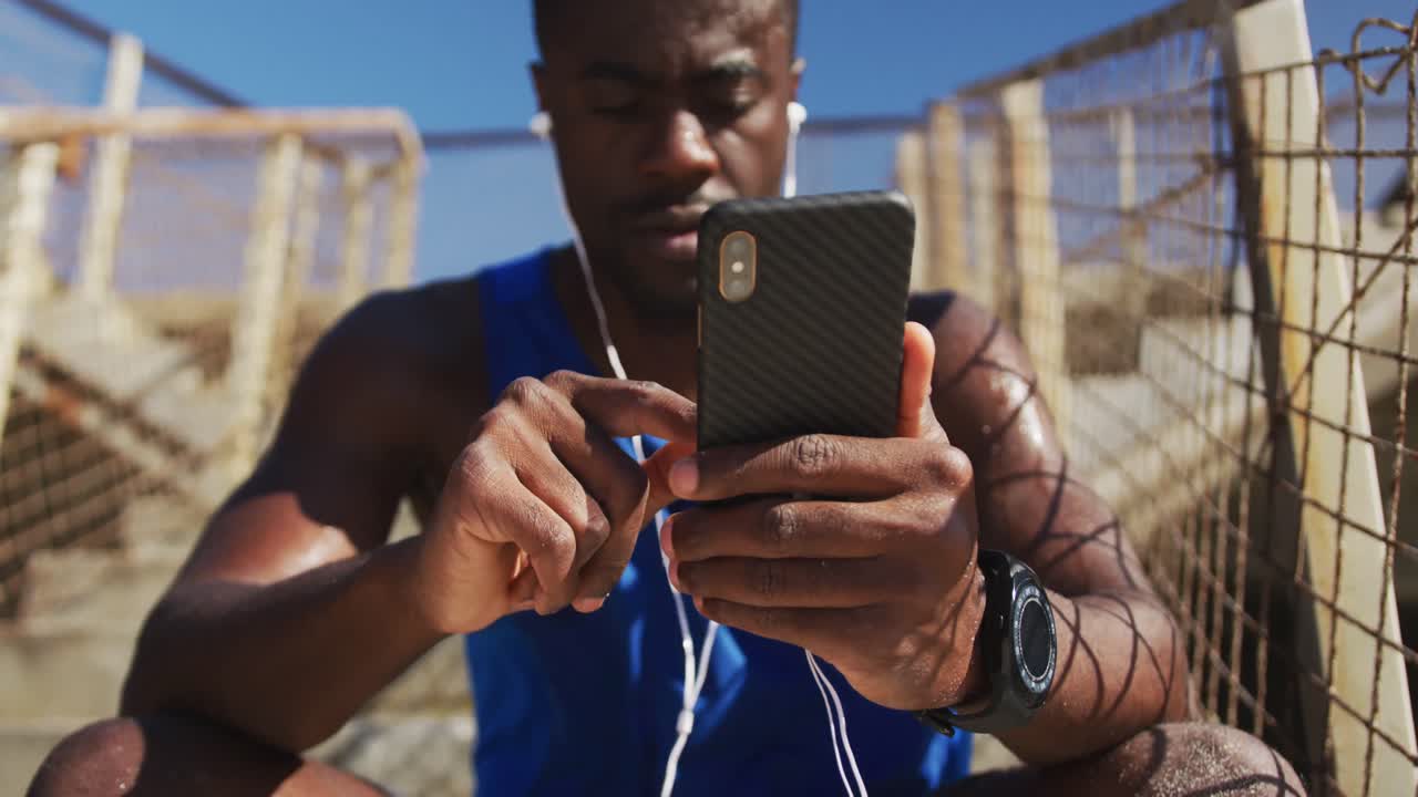 hombre afroamericano con un teléfono inteligente, tomando un descanso en el ejercicio al aire libre