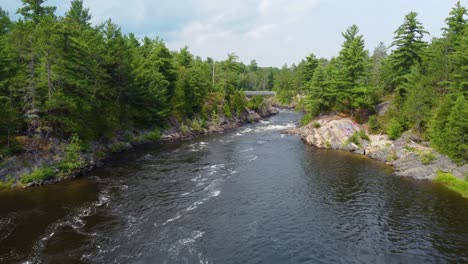 aerial dolly upsteam of rushing river with rocky shale banks and trees leading to car bridge