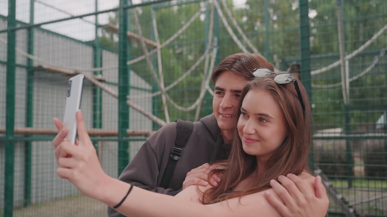 Coppia caucasica che posa per un selfie allo zoo, stretti in un abbraccio, content creator in stile influencer, estetica, fotocamera del telefono, fascia per capelli, occhiali da sole, sorrisi calorosi, momento romantico vicino a una staccionata verde con corda, esposizione diurna