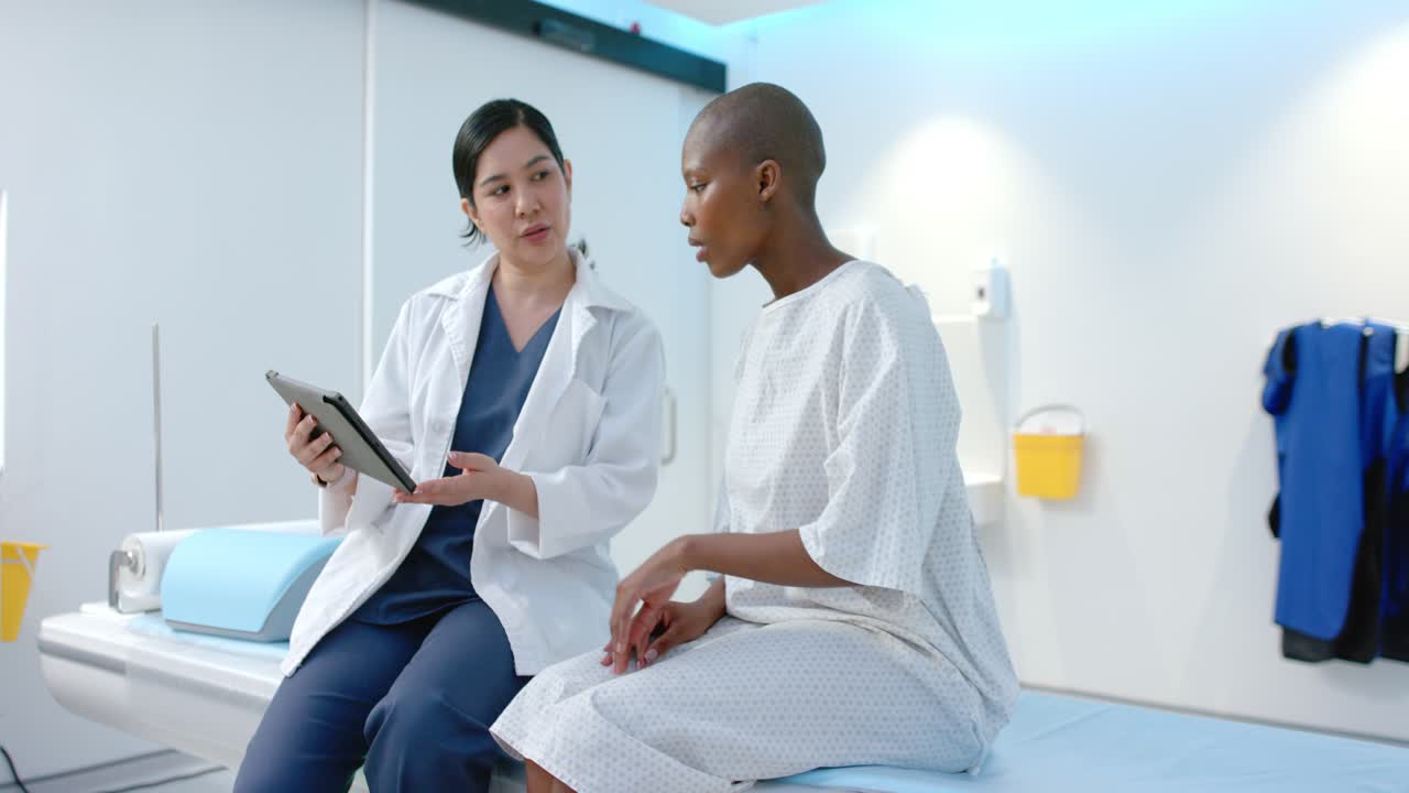 Doctor holding tablet explaining in exam room while African American patient in gown tapping screen