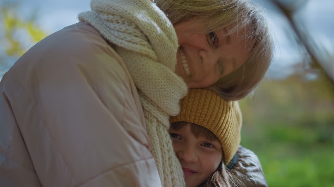 mom smiling joyfully while hugging daughter under golden autumn tree with leaf resting on head, capturing warmth, tenderness, and playful affection in cozy outdoor moment under soft natural light