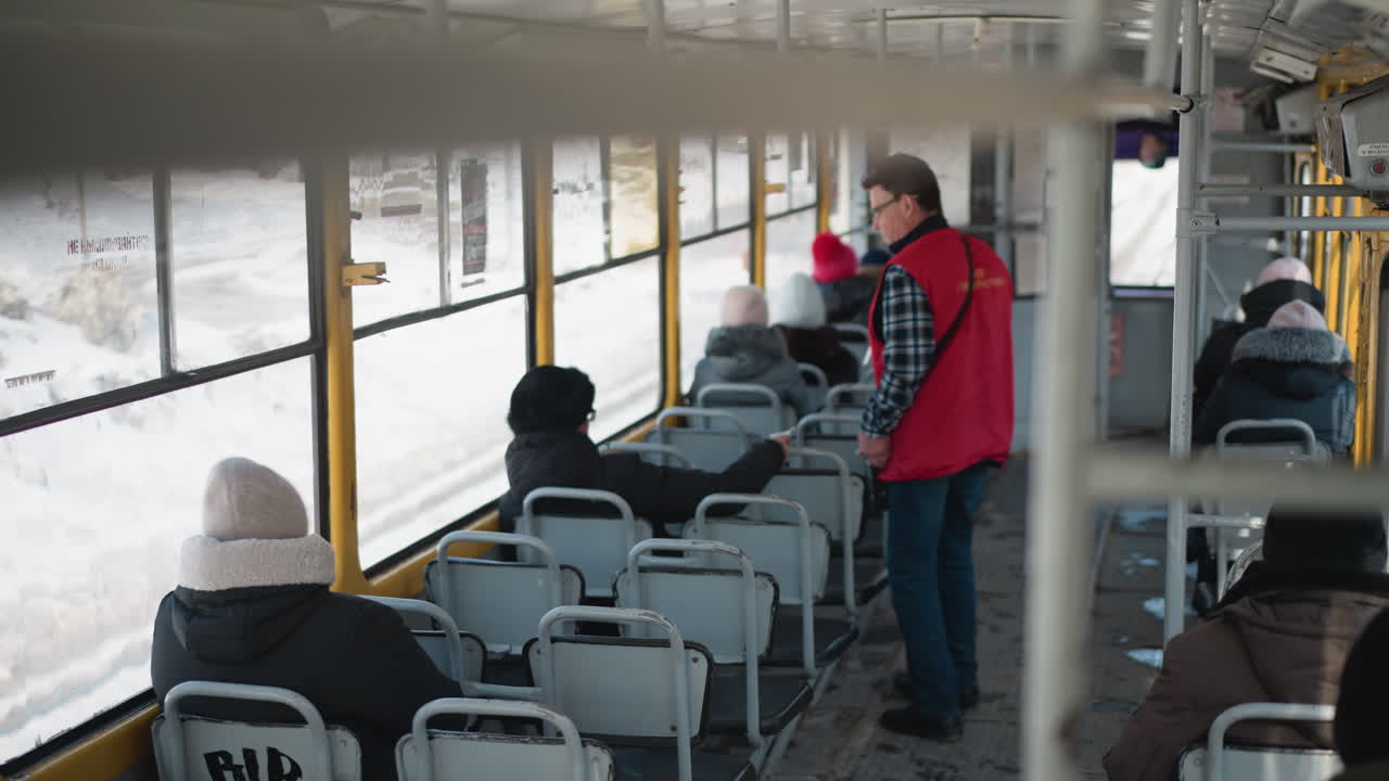 high angle view of ticket collector in red jacket with glasses moving forward through moving train interior to collect ticket from passenger, winter commute light on windows, calm urban transit mood