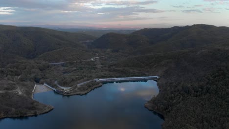 Fast-aerial-over-mountain-lake-dam-at-sunset-dense-forest-high-altitude-colorful