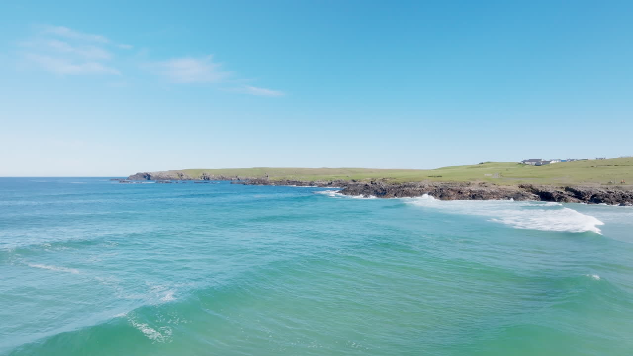 Aerial drone footage over Eoropie Beach on the Isle of Lewis, showing turquoise Atlantic water, reef-lined coast, and rolling green headlands near the Butt of Lewis. Clear blue skies and calm swell